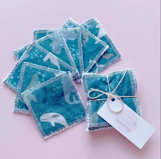 Image shows a pale pink background with one of small reusable wipes bundled with string and labelled with a rectangular tag next to pack of wipes fanned out to show a blue fabric featuring a print depicting whales and porpoises.