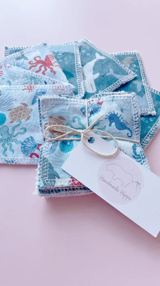 Image shows a pale pink background with one of small reusable wipes bundled with string and labelled with a rectangular tag next to pack of wipes fanned out showing two prints. One print is on blue fabric featuring a print depicting whales and porpoises, the other print is pale blue showing a range of sea creatures in blue and red