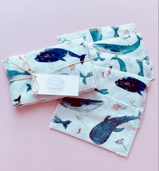 Image shows a pale pink background with one of small reusable wipes bundled with string and labelled with a rectangular tag next to pack of wipes fanned out to show a pale blue fabric featuring a print depicting multiple types of sea creature.