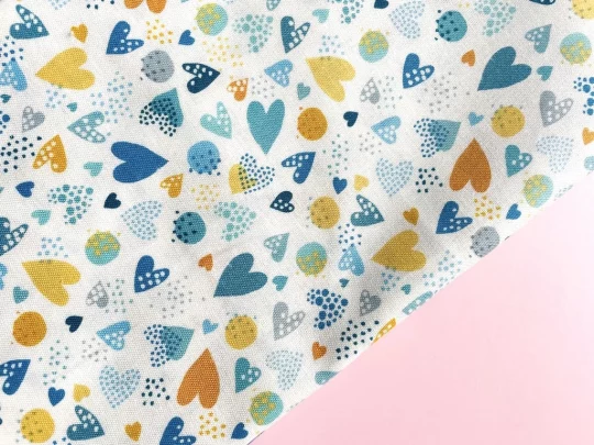 Piece of fabric on pink backdrop. Fabric is white with hearts and circles of different sizes in various shades of yellow and blue