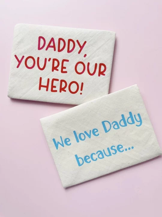 two closed finished envelopes, slightly overlapped on pink background. One envelope says "Daddy, you're our hero!" in red capital letters and the other says "We love Daddy because..." in light blue text.
