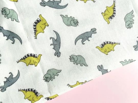 Piece of fabric on pink backdrop. Fabric is off-white with a scattered dinosaur design showing stegosaurs, triceratopses and T-Rexes in various shades of green.