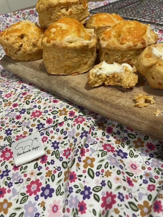 Image shows a pile of cheese scones on a wooden chopping board, one scone has been split in half, buttered and a bite has been taken. The chopping board is placed on top of a floral apron