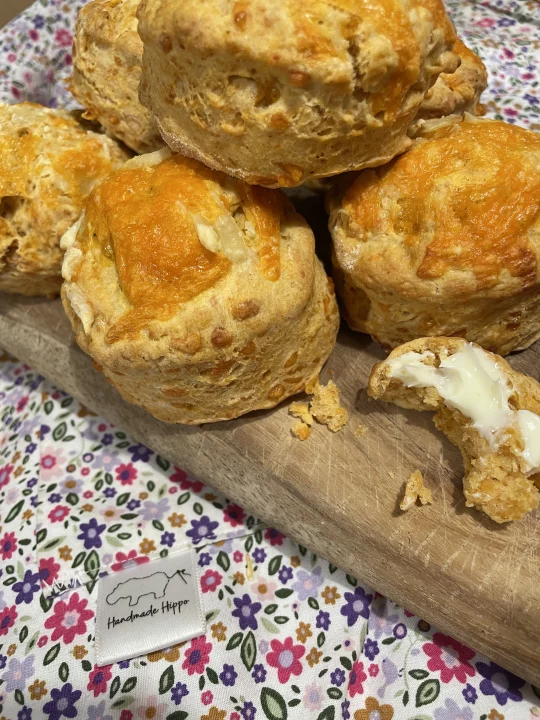 Image shows a pile of cheese scones on a wooden chopping board, one scone has been split in half, buttered and a bite has been taken. The chopping board is placed on top of a floral apron