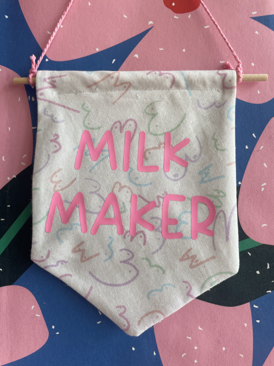 Image shows small cotton banner hanging by a pink thread on a pink and blue floral background. The banner is made from white fabric with a multiple line drawings of boobs in different shapes and sizes in a pastel colour palette. Pink text on the banner reads "MILK MAKER"