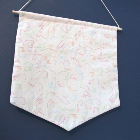 Image shows large cotton banner hanging by a white thread on a dark blue background. The banner is made from white fabric with a multiple line drawings of boobs in different shapes and sizes in a pastel colour palette.