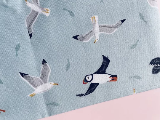 Piece of fabric on pink backdrop. Fabric is pale grey-blue with flying puffins and gulls with the silhouettes of some fish scattered between the birds