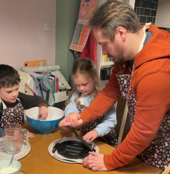 image shows a father and 2 young children preparing food together around a kitchen table, they are all wearing matching aprons