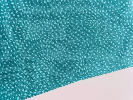 Piece of fabric on pink backdrop. Fabric is teal with small irregular pale blue dots in a interlinking arches design